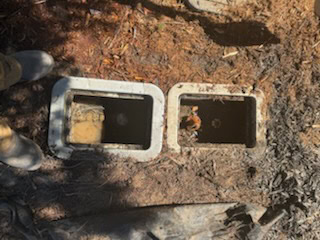 Two rectangular utility boxes set in the ground, one filled with muddy water and the other with dry leaves, surrounded by dirt and dry grass. A plumber El Dorado County, CA is partially visible in boots at the lower left corner.