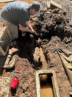 A plumber El Dorado County, wearing a cap and dark shirt, is repairing a muddy, partially flooded pipe in a trench, surrounded by tools and wet soil.