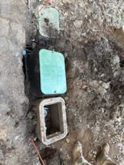 A green utility box and an empty concrete box are set in the ground surrounded by dirt and gravel, with some red markers nearby—a common sight for a plumber El Dorado County might encounter during water heater replacement work.