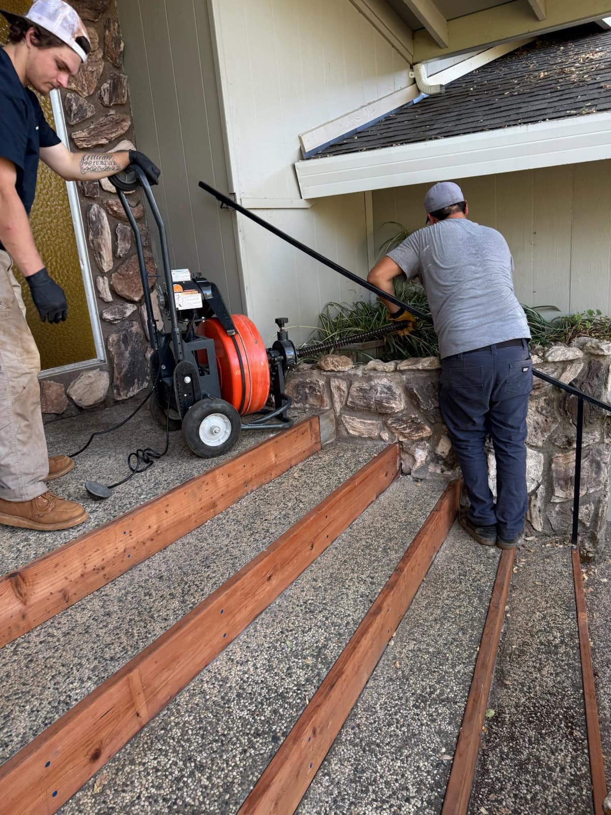 Two men, including a plumber El Dorado County, use a drain cleaning machine on a stone-lined porch. One feeds the cable into the drain while the other operates the equipment near wooden stair railings by the home's entrance.