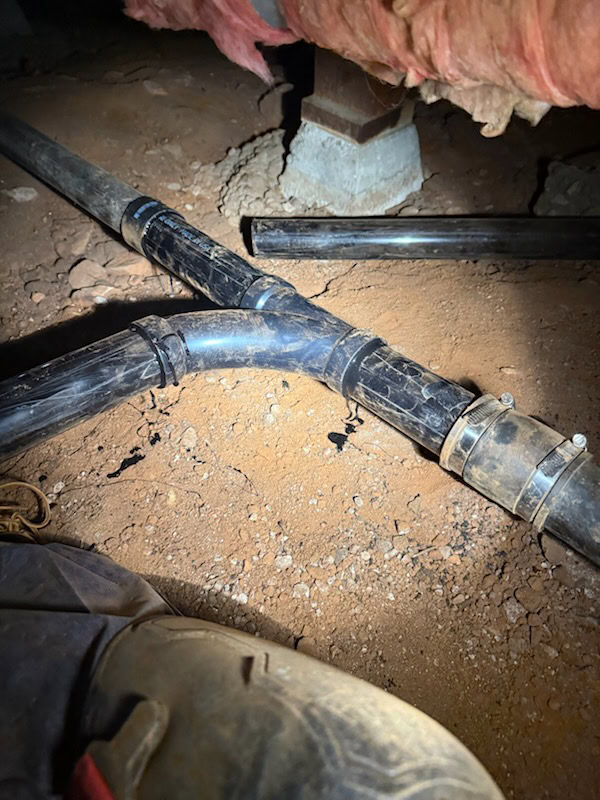 A close-up of black plumbing pipes joined with metal clamps, laying on reddish dirt under a CA house with exposed insulation—a common scene during water heater replacement El Dorado County plumbers handle.