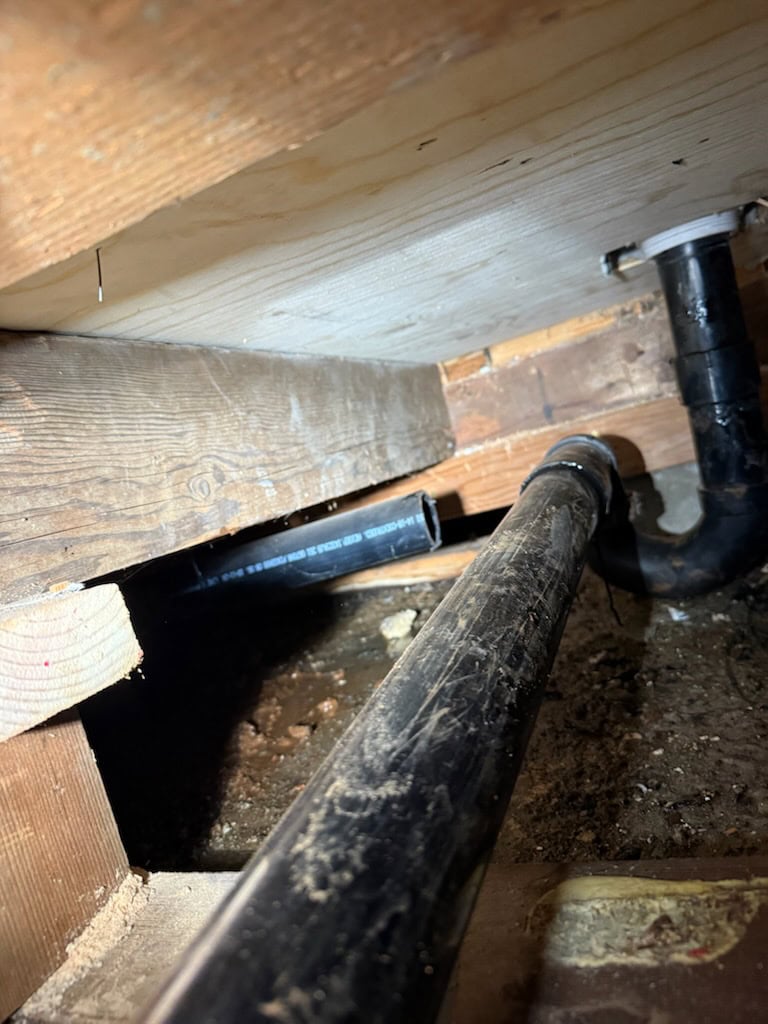 A close-up view of black PVC pipes under a wooden floor, with one pipe disconnected, surrounded by dirt and wooden beams in a crawl space—an issue best handled by a skilled plumber El Dorado County.