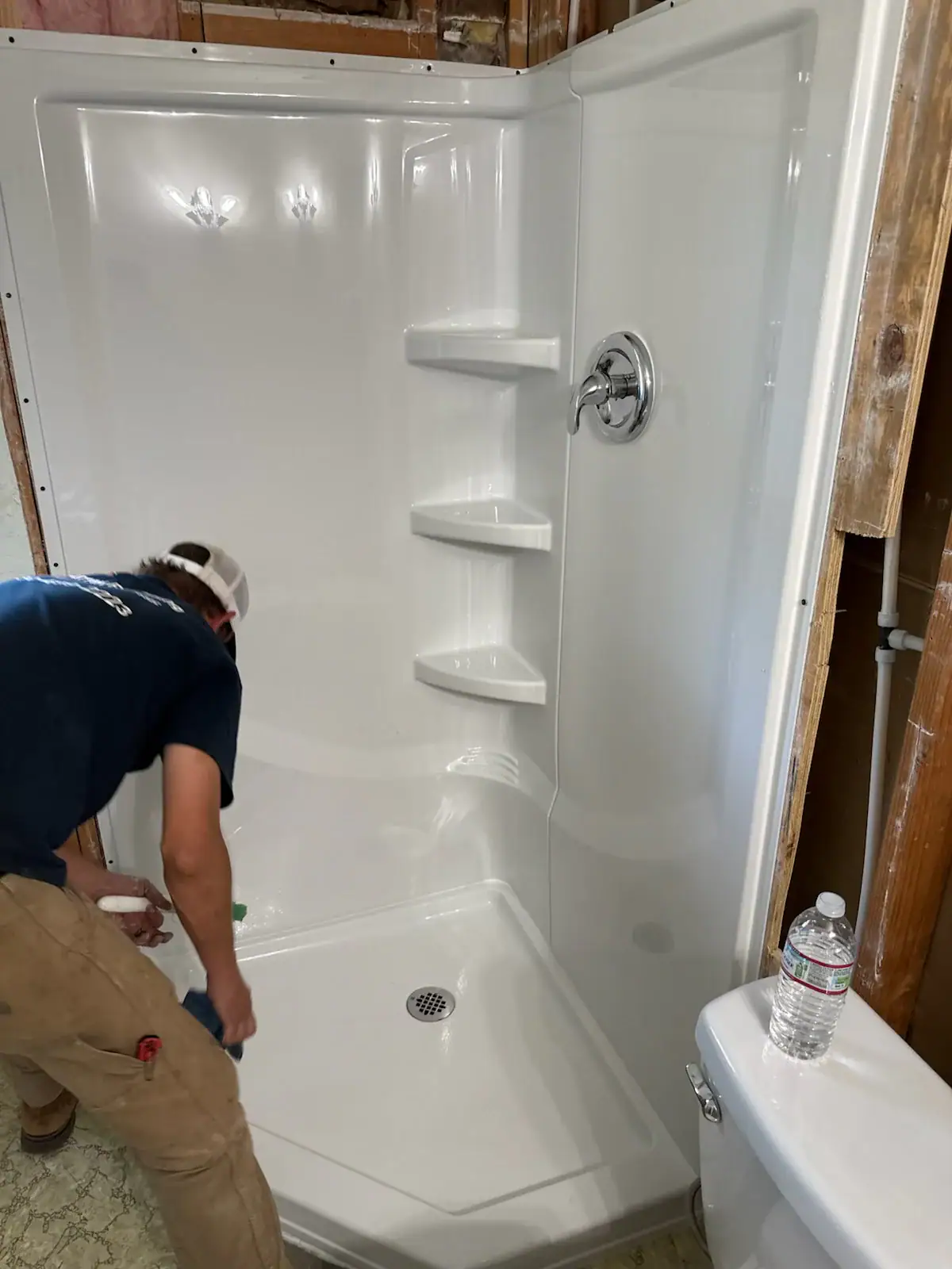 A person in a blue shirt and tan pants cleans a newly installed white shower stall with corner shelves and a chrome shower handle. Exposed wooden wall studs and a water bottle on a toilet are visible nearby.