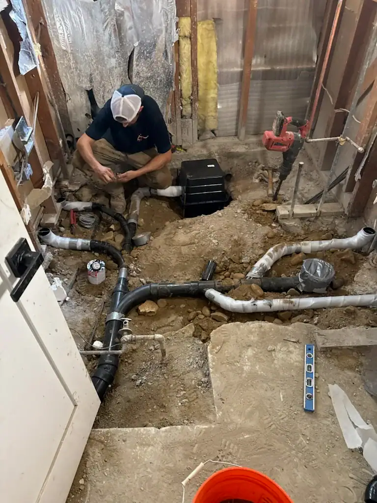 A plumber kneels in a partially demolished bathroom, working on exposed black PVC pipes and drains in the floor. Tools, a level, and construction materials are scattered around the unfinished space.