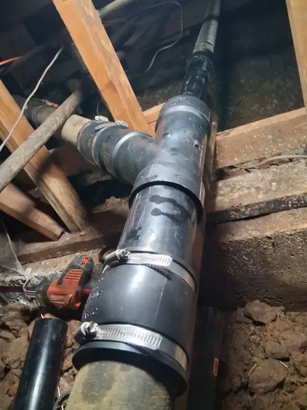 A close-up view of black PVC pipes with metal clamps and fittings installed in a crawl space with exposed wooden beams and dirt floor. A power tool is visible to the left.