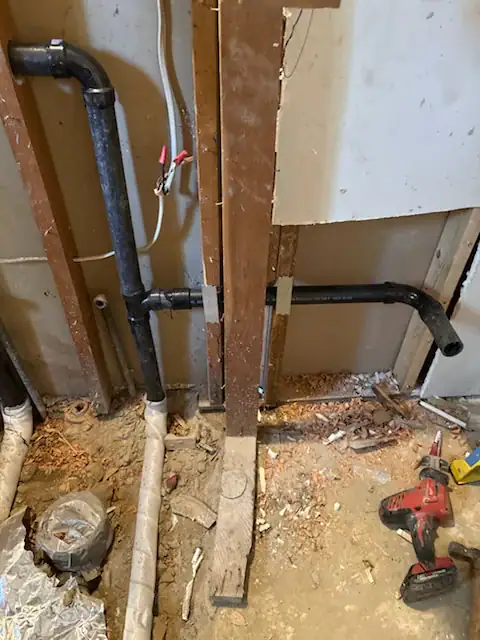 A partially demolished wall reveals exposed black plumbing pipes, electrical wiring, wood framing, construction debris, and a red cordless power drill lying on the dusty floor.
