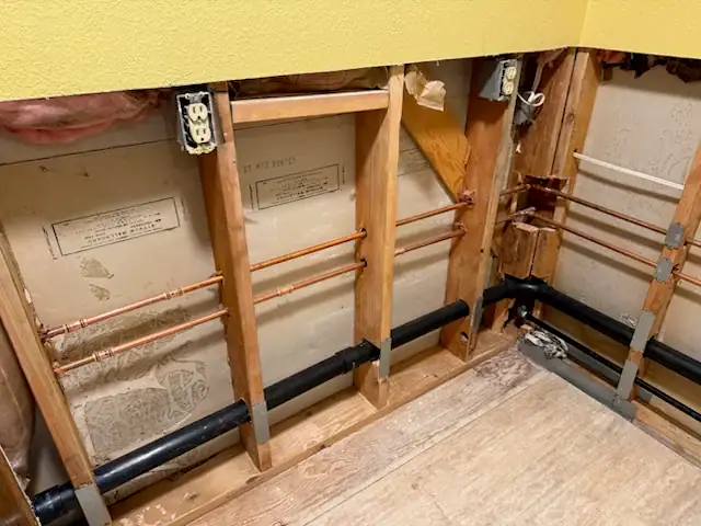 Exposed wall studs with visible plumbing pipes, electrical outlets, and insulation in a partially renovated room with a plywood floor and yellow-painted upper wall.
