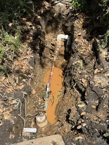 A muddy trench with exposed water pipes and valves, partially filled with murky brown water, surrounded by soil, roots, and scattered leaves.