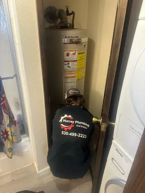 A plumber in a black Murray Plumbing jacket kneels in front of a water heater inside a small closet, working. A stacked washer and dryer are to the right, and part of a bathroom with a shower is visible on the left.