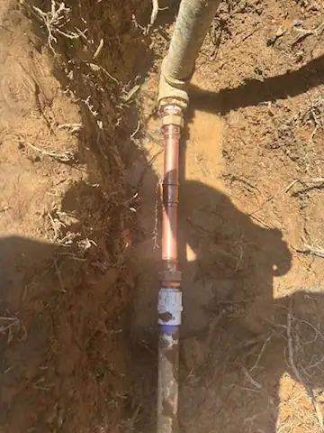 A section of copper pipe with fittings is installed in a trench with exposed soil and roots, connecting to two other pipes. Shadows partially cover the area.