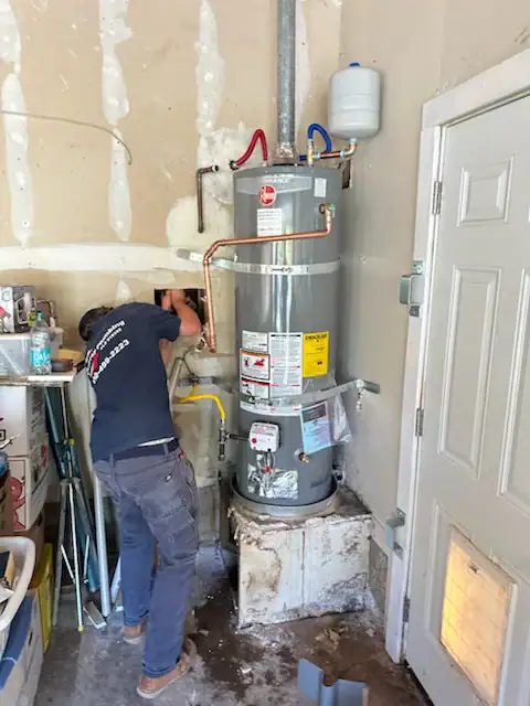 A person is installing or repairing a water heater in a garage, standing next to the unit with tools in hand. The water heater is elevated on a platform, with visible pipes and warning labels.