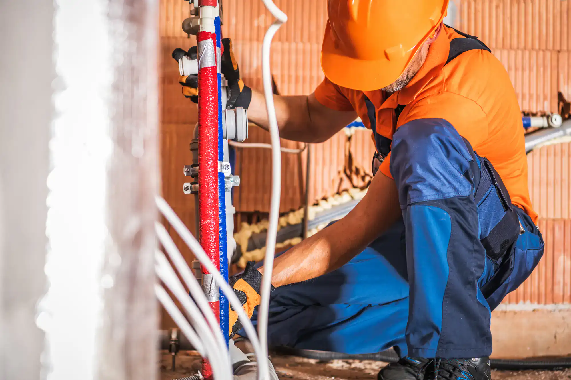 A construction worker in an orange hard hat and safety gear installs or repairs plumbing pipes inside a building, using tools and focusing on a blue and red pipe system.