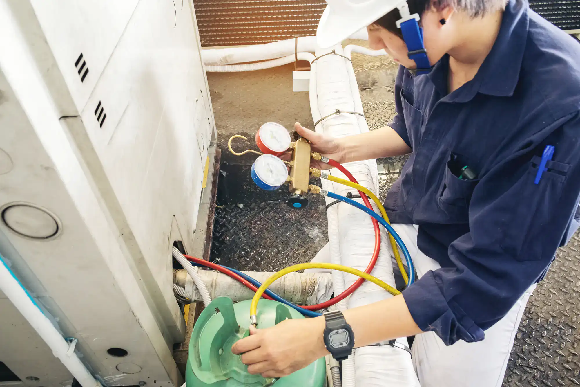 A technician wearing a white hard hat and navy uniform checks pressure gauges on an HVAC system, using colored hoses connected to a green refrigerant tank.