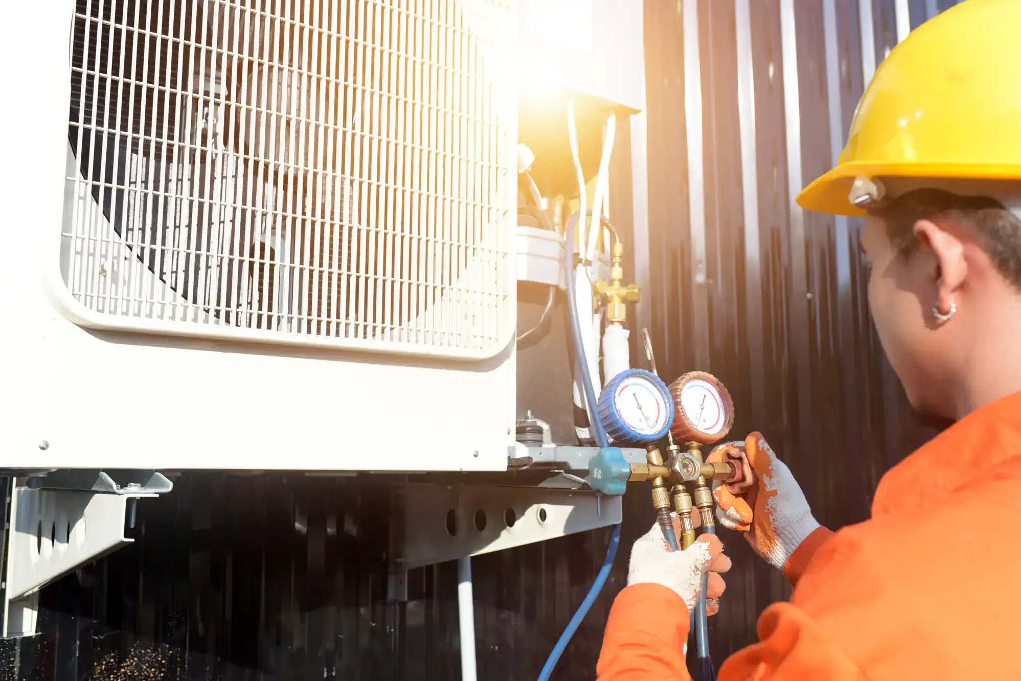 A technician wearing a yellow hard hat and orange safety uniform uses a manifold gauge to check an outdoor air conditioning unit in bright sunlight.