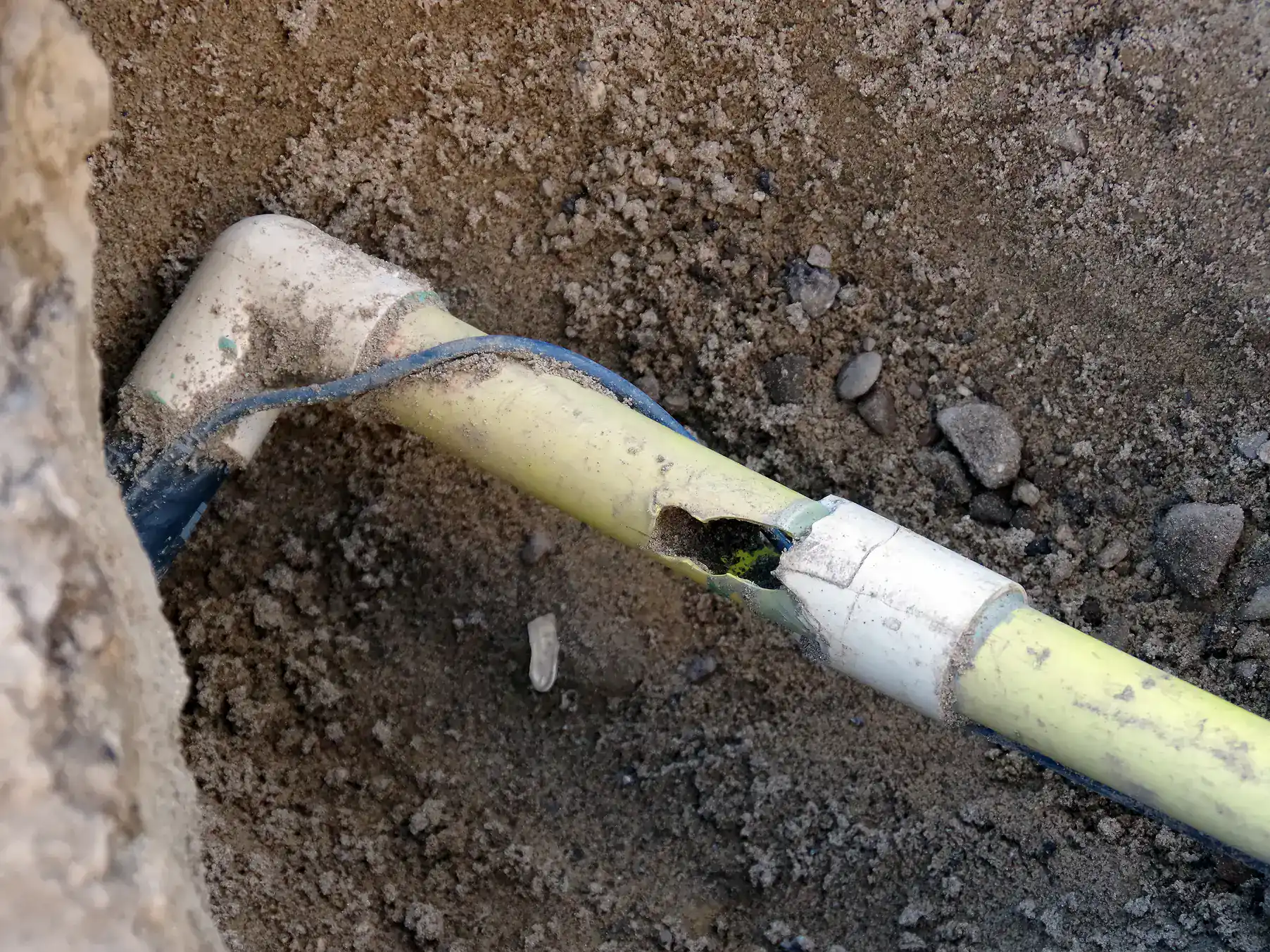 A close-up of a broken plastic pipe underground, showing a crack and damage, surrounded by soil and small rocks.