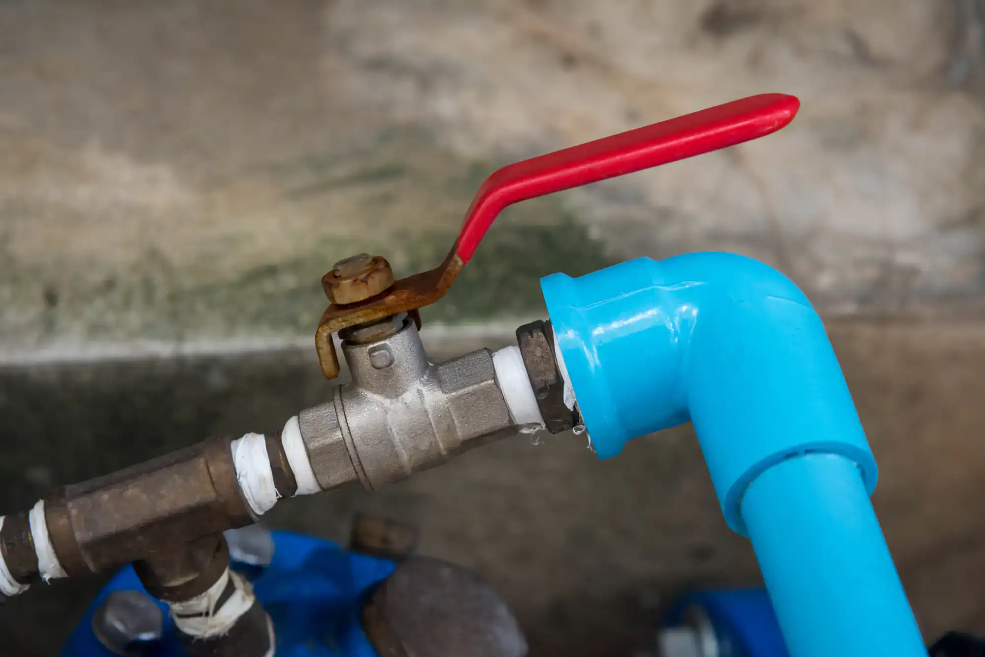Close-up of a metal water valve with a red lever handle connected to blue PVC pipes, attached to a wall with visible signs of wear and slight water leakage.