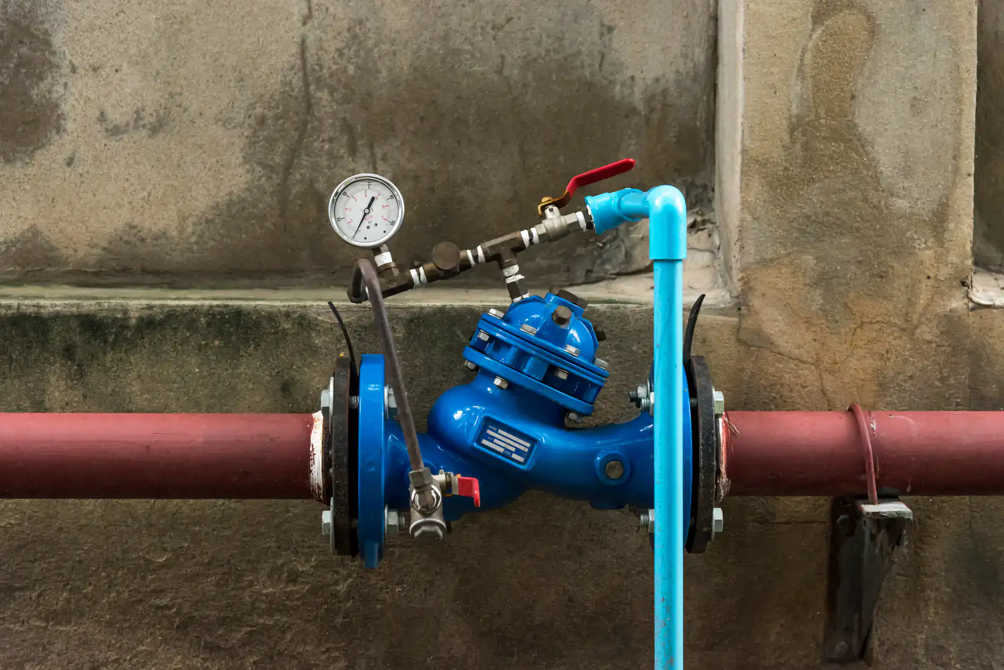 A blue water pressure valve with a gauge and red-handled lever is connected to horizontal red pipes and a vertical blue pipe, mounted against a weathered concrete wall.
