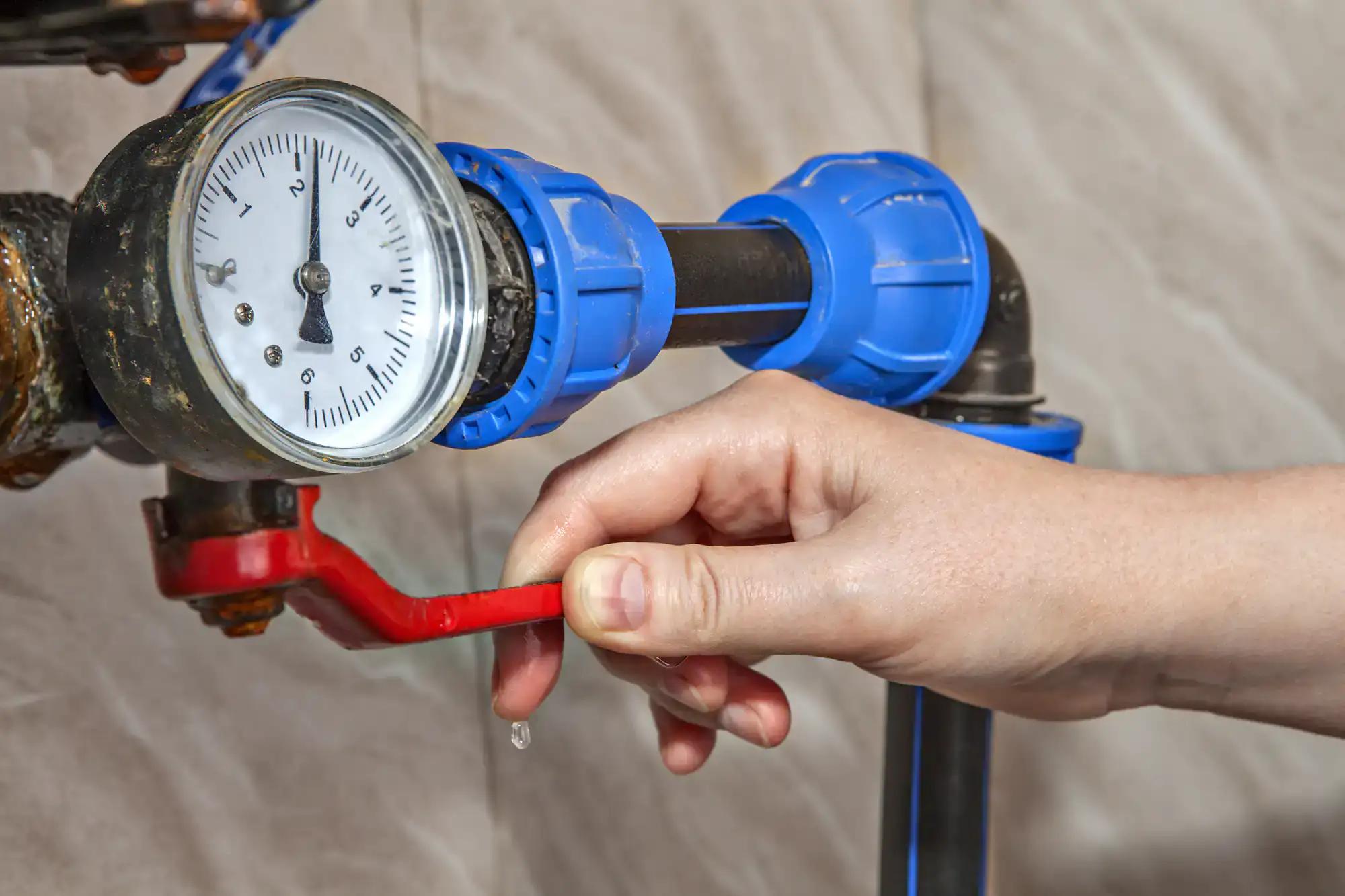 A hand turns a red valve on a black pipe connected to a pressure gauge, with blue pipe fittings. A water droplet is visible near the valve.