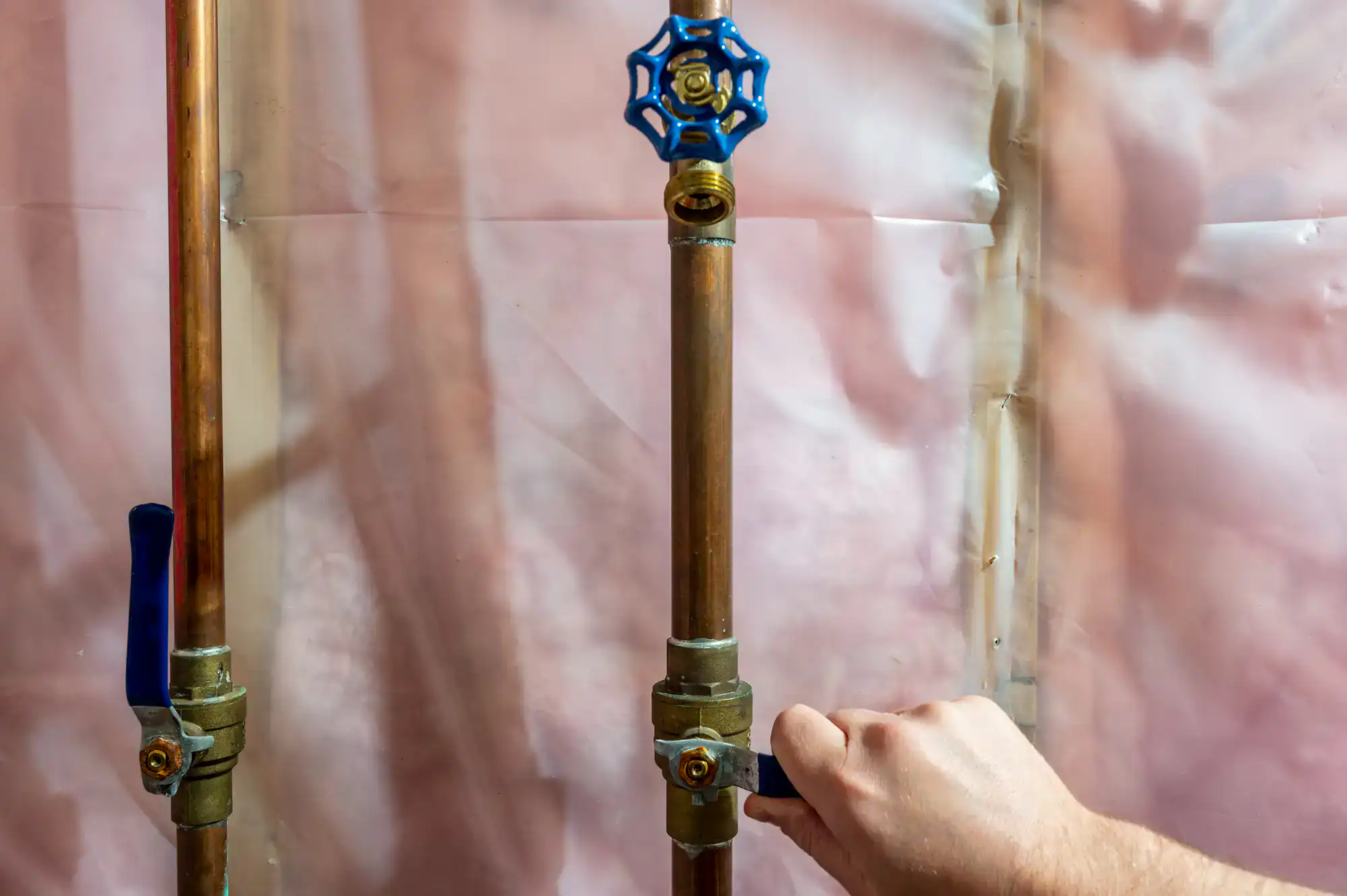 A hand turns the blue handle of a shut-off valve on a copper water pipe against a wall covered in pink plastic sheeting.