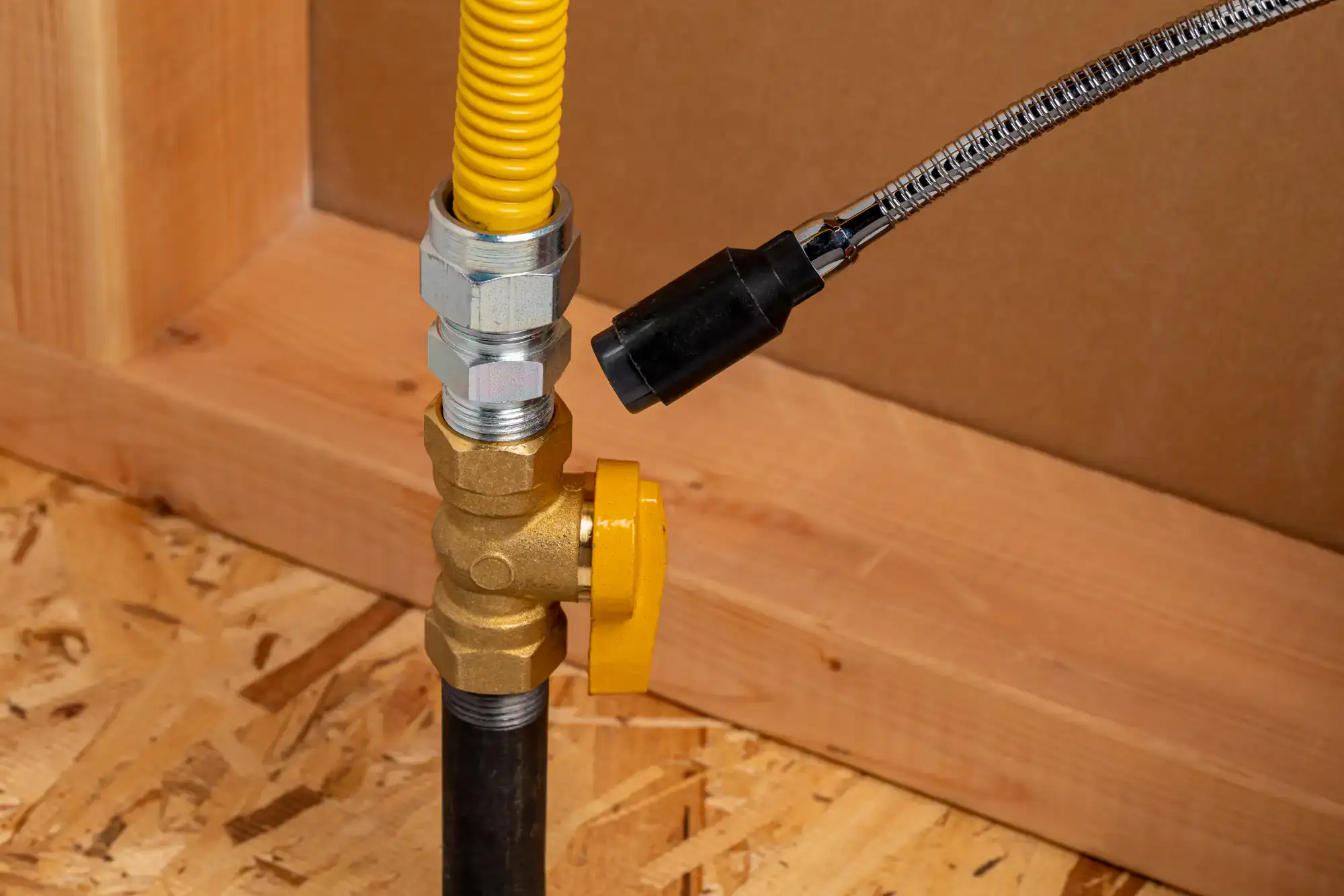 Close-up of a gas valve with a yellow handle, connected to a black pipe and flexible yellow and silver hoses, mounted on a wooden board background.