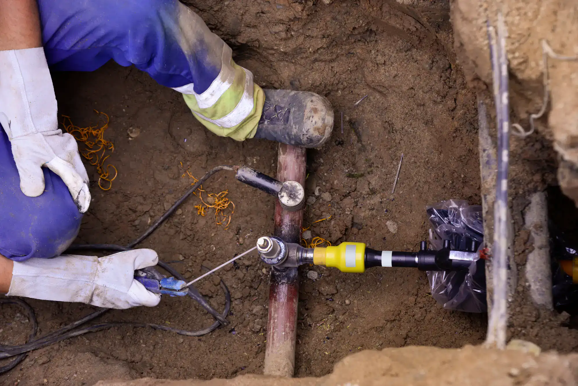 A worker wearing gloves and blue pants repairs a buried pipe using tools and equipment in a trench dug into the soil.