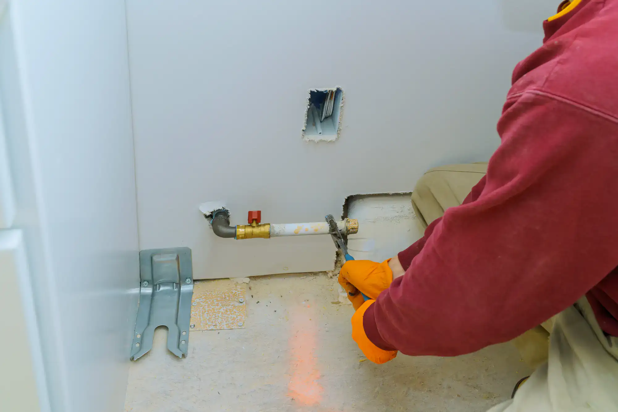 A person wearing orange gloves and a red shirt works on a white pipe coming out of a wall, possibly performing plumbing repairs. The wall has two cutouts and construction materials are visible on the floor.