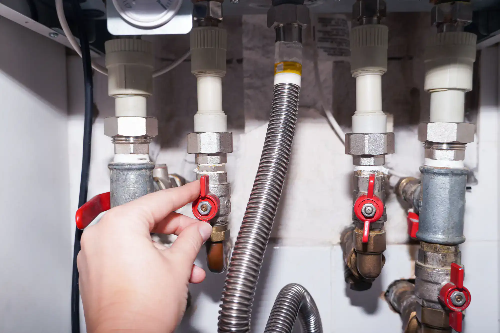A hand turning a red valve on a metal pipe among several pipes and fittings connected to plumbing or heating equipment.