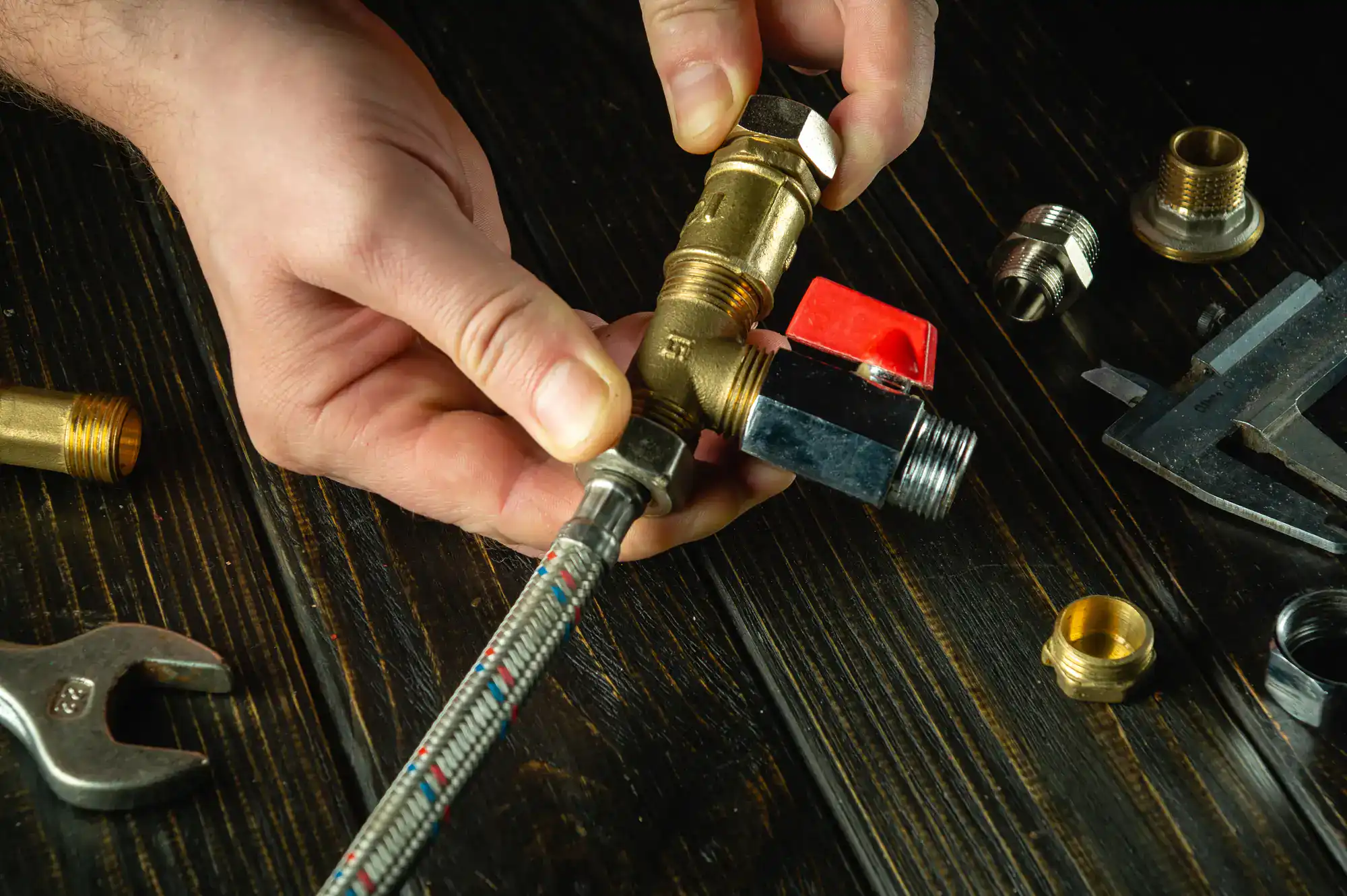 A person’s hands assembling metal plumbing fittings and a flexible hose on a dark wooden surface, surrounded by various plumbing tools and parts.