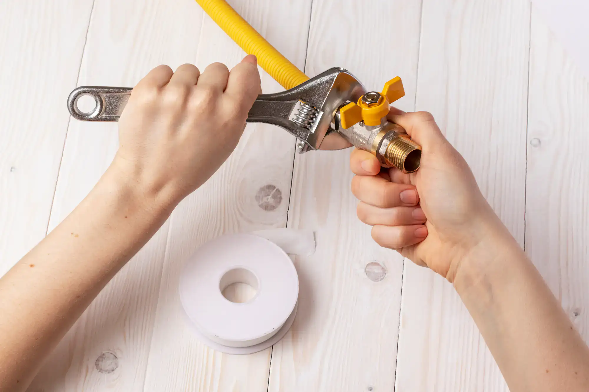 A person uses a wrench to tighten a yellow gas valve, while holding it steady with the other hand. A roll of white plumber’s tape lies on a light wooden surface nearby.