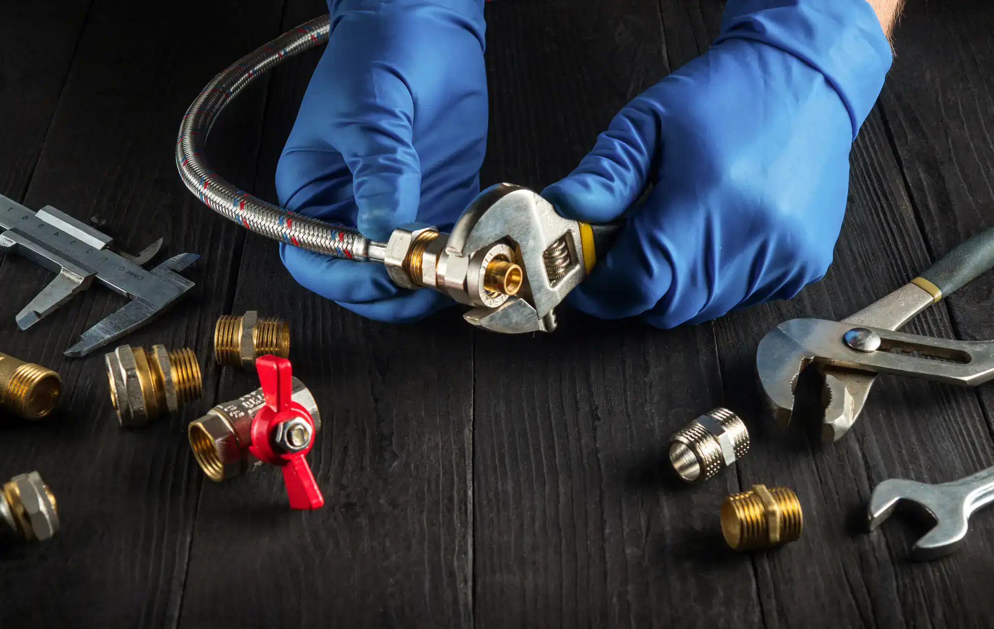 A person wearing blue gloves uses a wrench to tighten a metal hose connection on a dark wooden surface, surrounded by various brass fittings, a valve, and tools.
