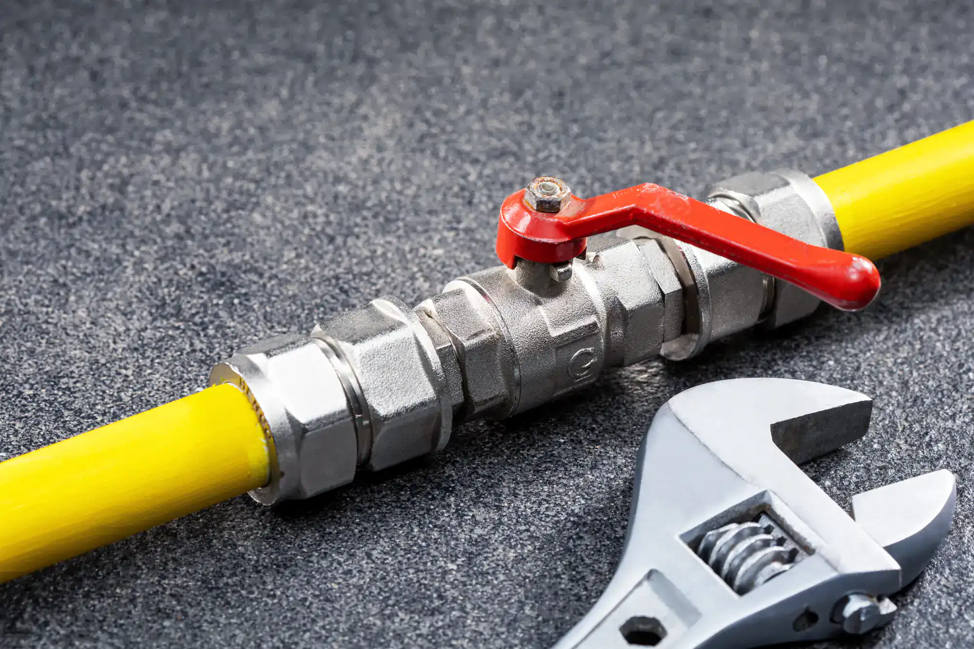 A yellow gas pipe with a metal shutoff valve featuring a red lever handle is lying on a gray surface, next to a silver adjustable wrench.