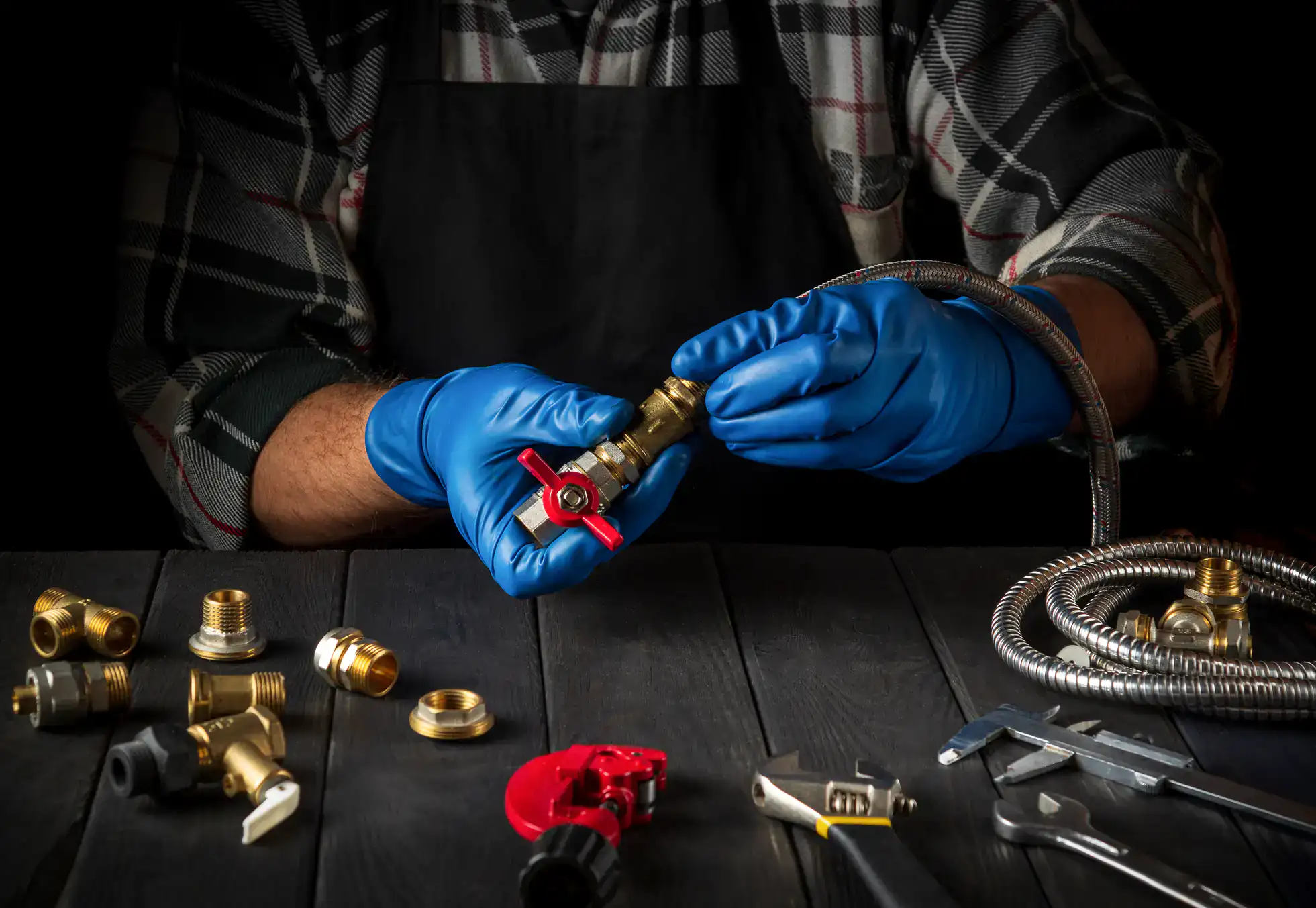 A person wearing blue gloves and a plaid shirt works with brass plumbing fittings and flexible hose at a dark wooden table, surrounded by various plumbing tools and components.