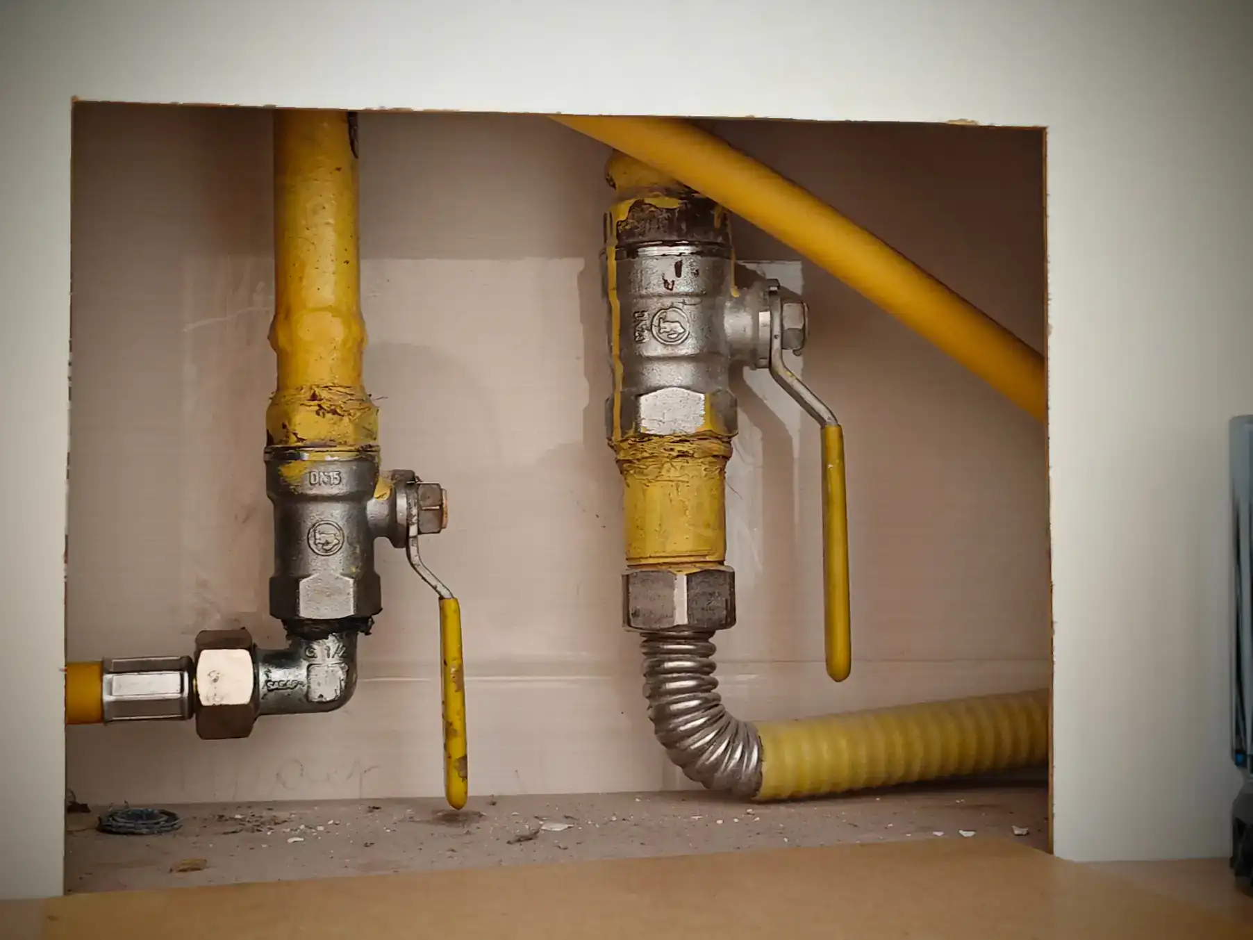 Two yellow gas pipes with metal valves and handles are installed through a rectangular opening in a wall. The pipes and valves show signs of wear and some corrosion.