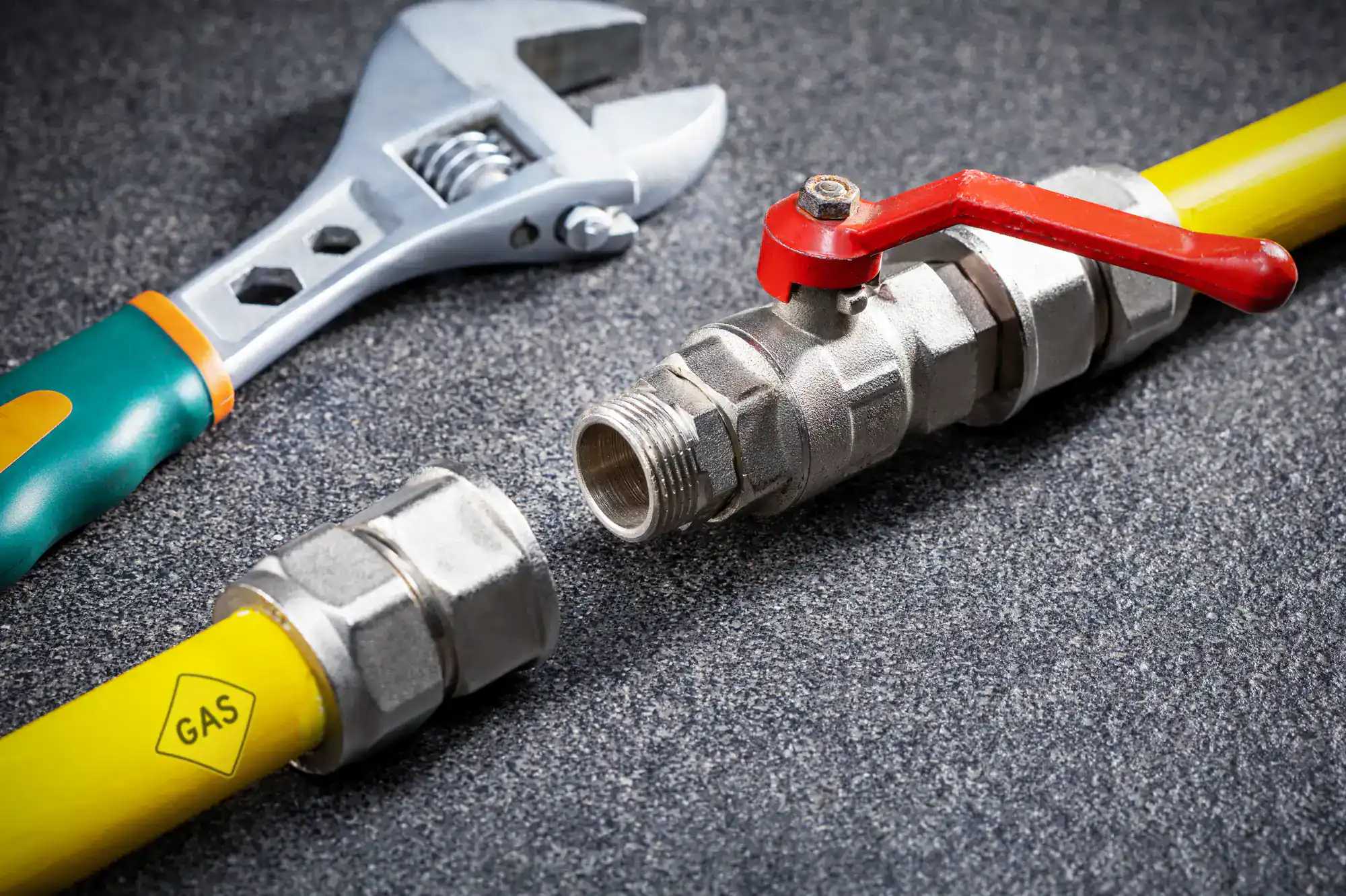 A gas pipe with a valve and a wrench on a textured gray surface. The pipe is disconnected, with visible threads, and the yellow pipe is labeled "GAS.