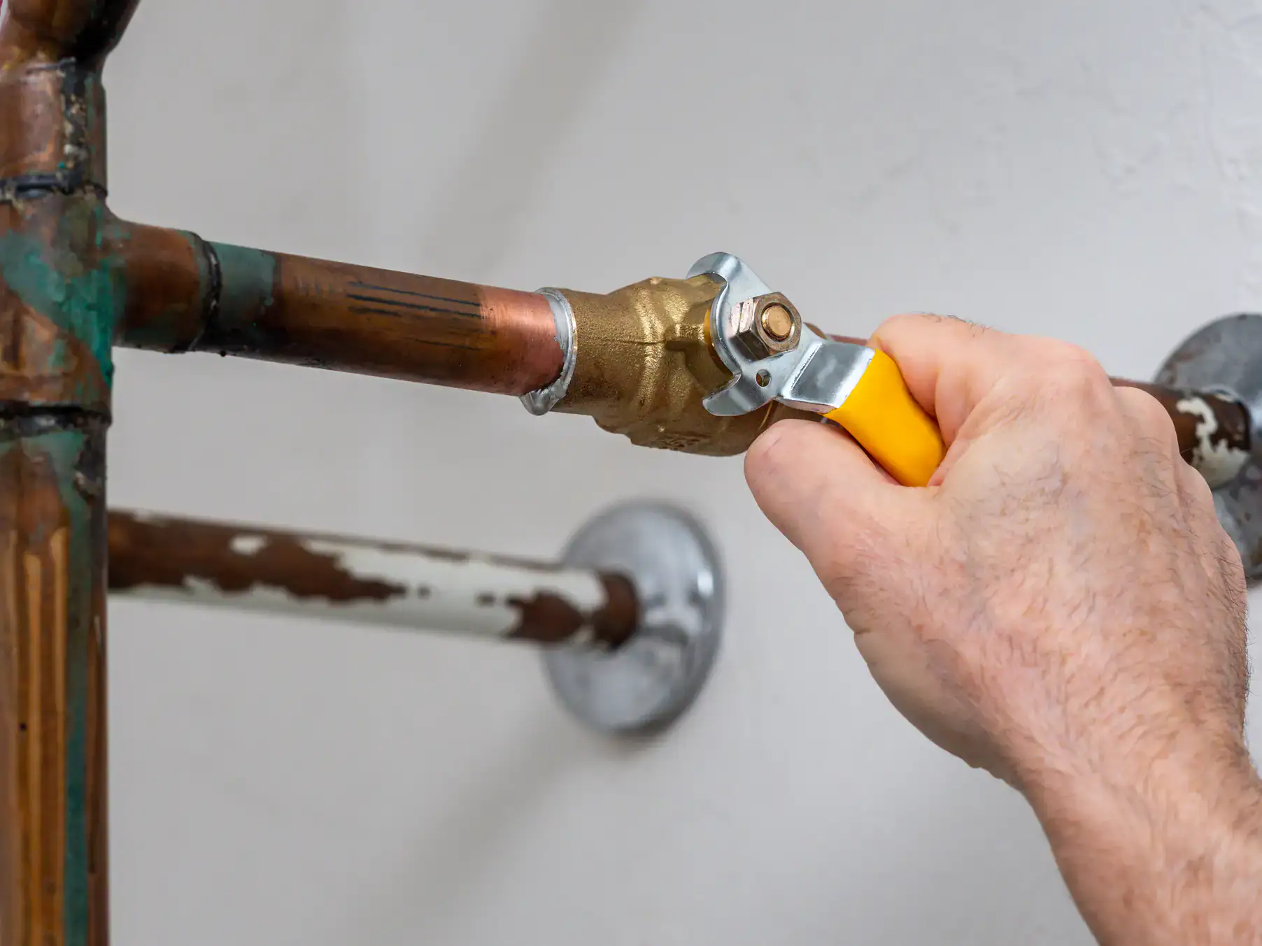 A close-up of a person’s hand turning a yellow-handled shut-off valve on a copper water pipe, with other pipes and a wall visible in the background.