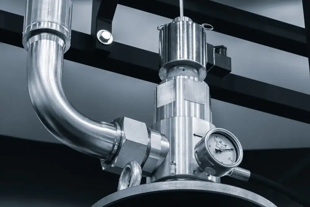 Close-up of industrial stainless steel piping with a pressure gauge attached, showing a reading of just above zero. The setup is part of a larger mechanical or manufacturing system.