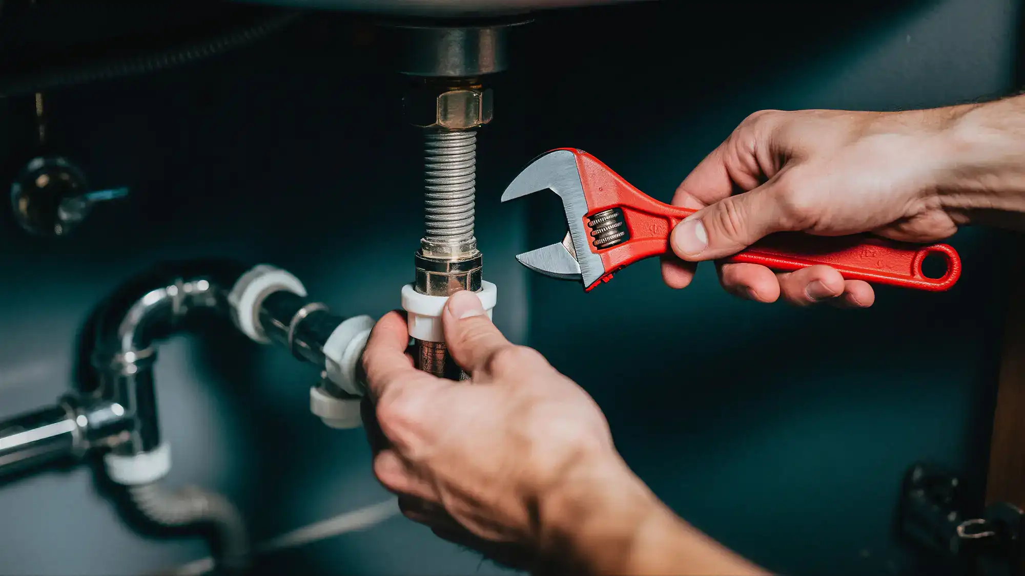 A person uses a red adjustable wrench to tighten a metal pipe under a sink, focusing on plumbing work with visible pipes and fittings.