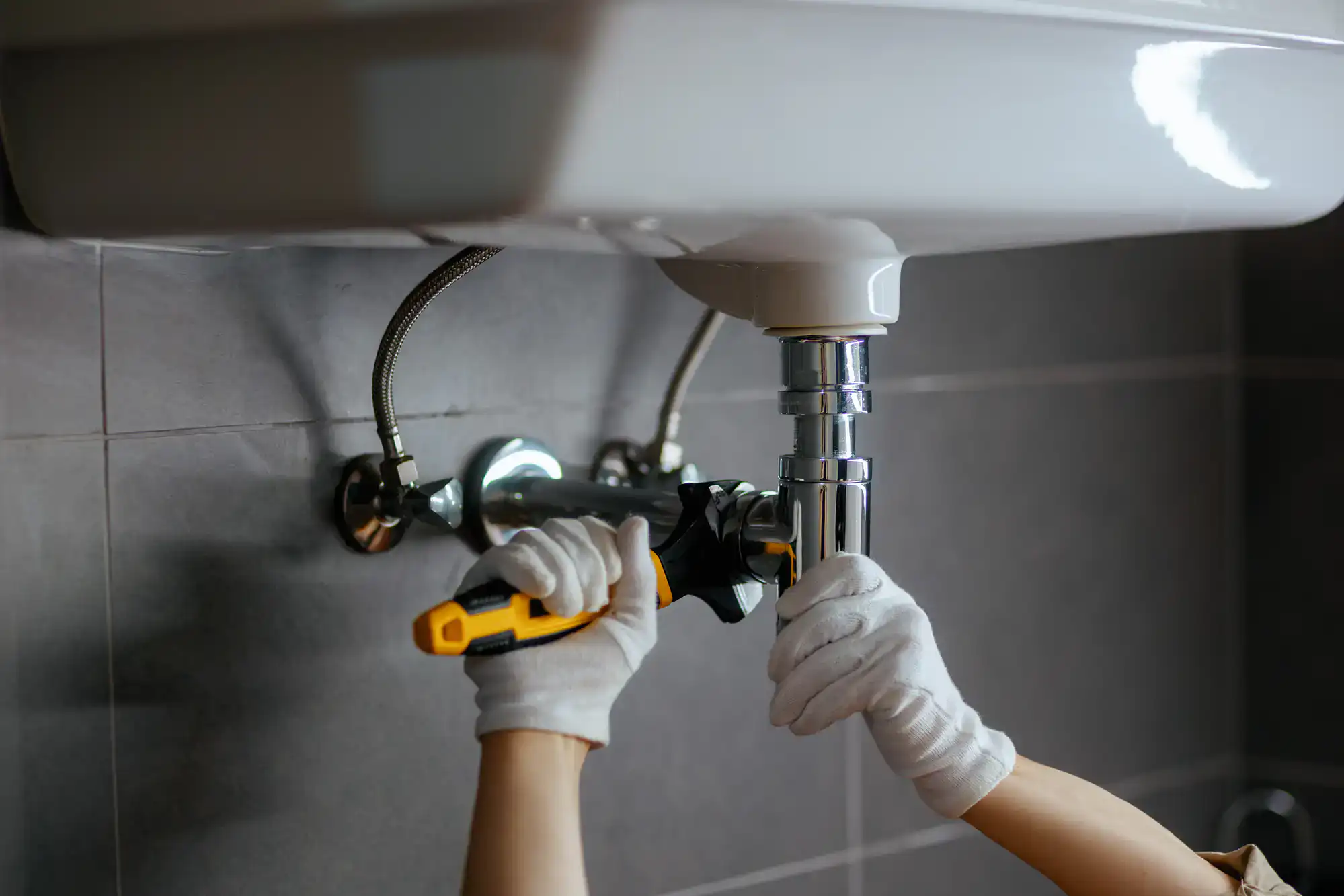 A person wearing white gloves uses a wrench to tighten a metal pipe under a white bathroom sink, performing plumbing repairs against a tiled wall.