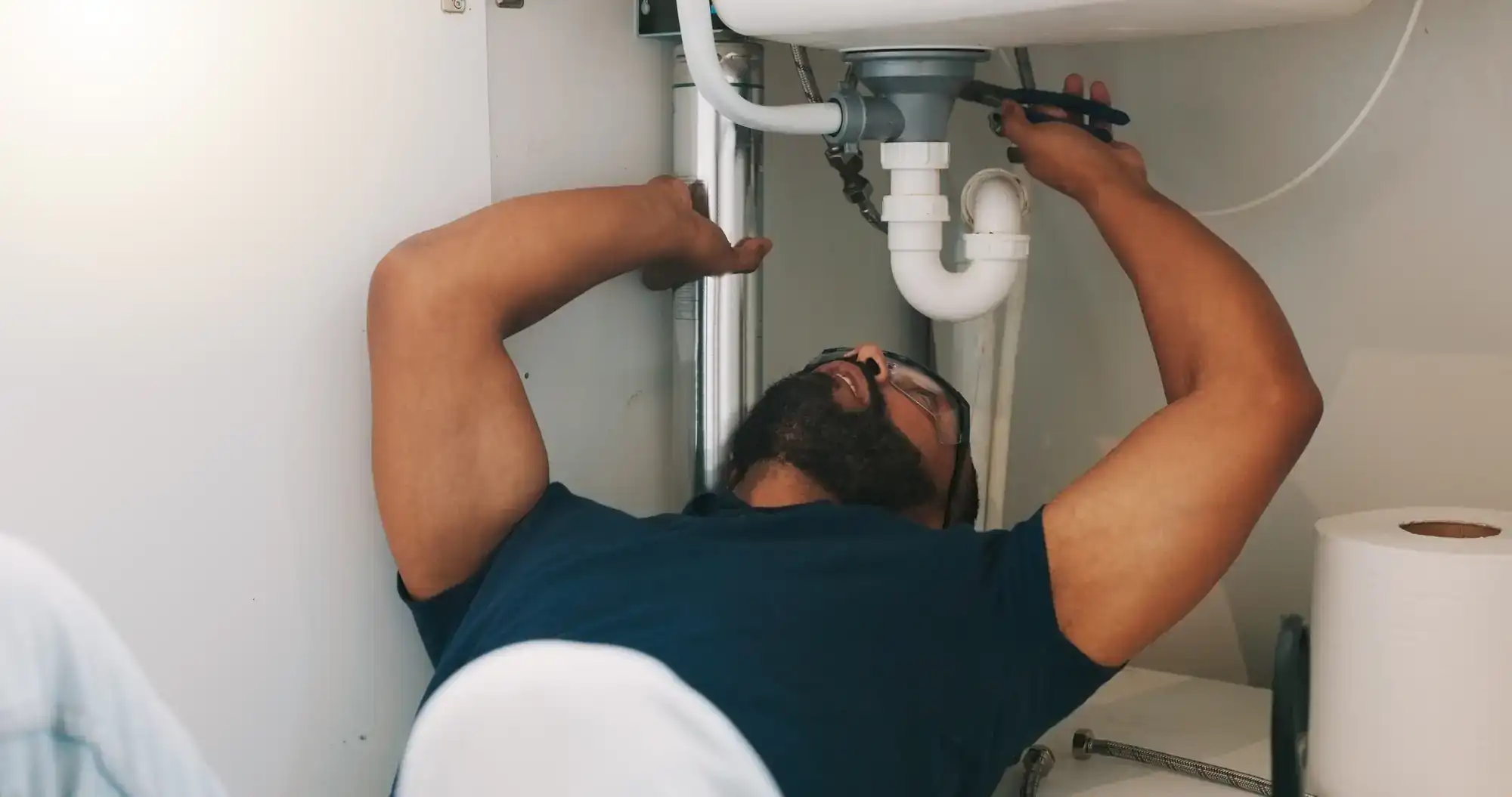 A man lies on his back under a sink, using a wrench to fix plumbing pipes. He is wearing glasses and a navy blue shirt, with tools and a roll of paper towel nearby.