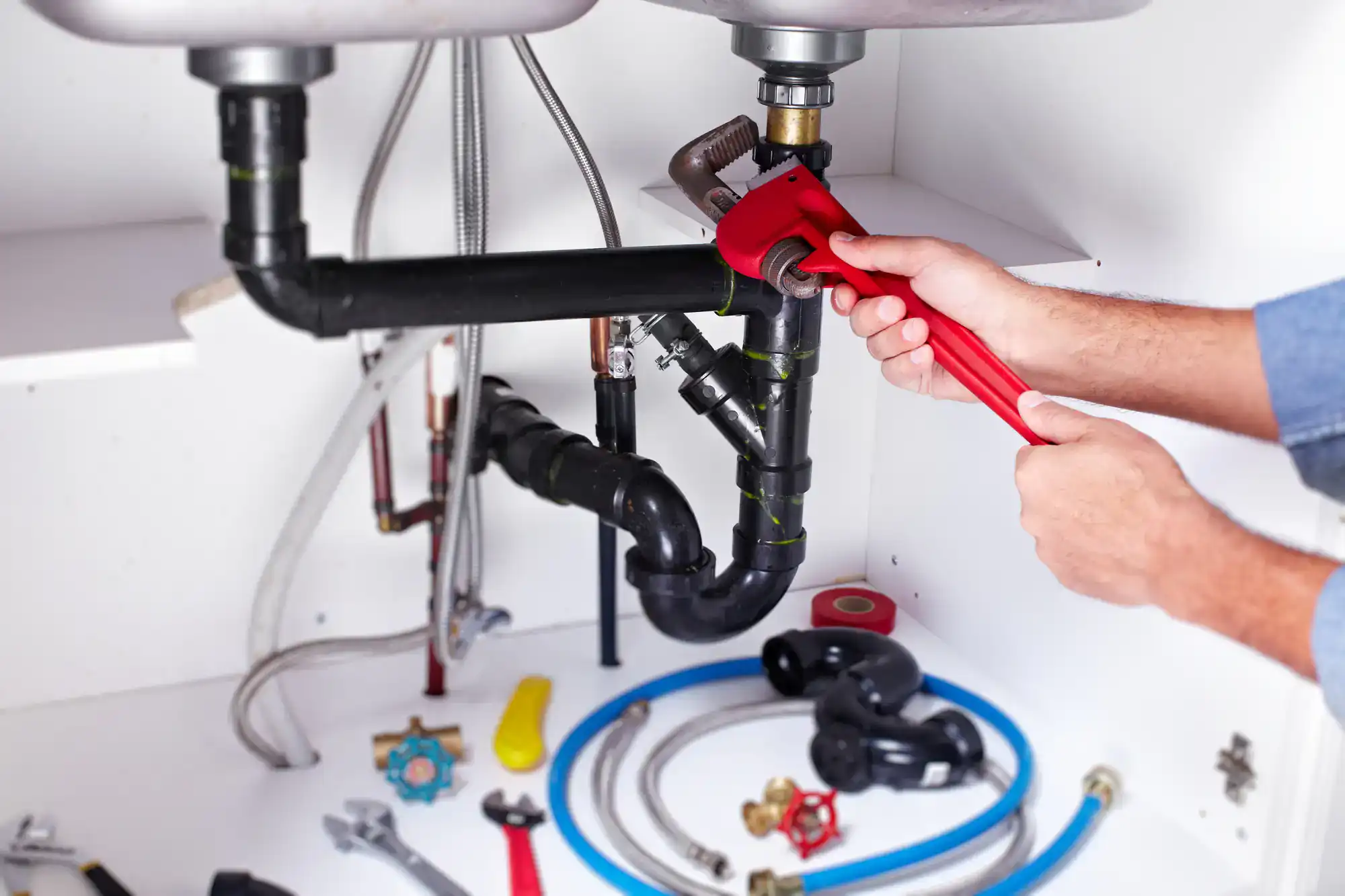 A person uses a red pipe wrench to tighten a pipe under a sink; various plumbing tools and supplies are spread out on the cabinet floor.