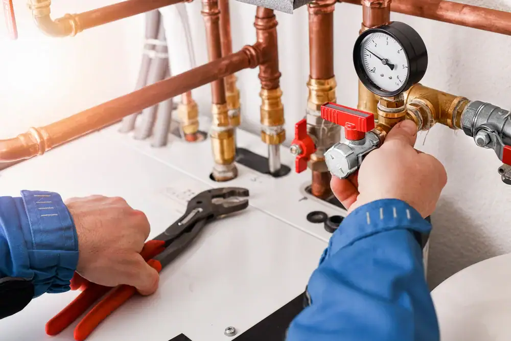 A person in blue work clothes adjusts a valve on a heating system or boiler, surrounded by copper pipes and a pressure gauge, with pliers and a wrench in hand.