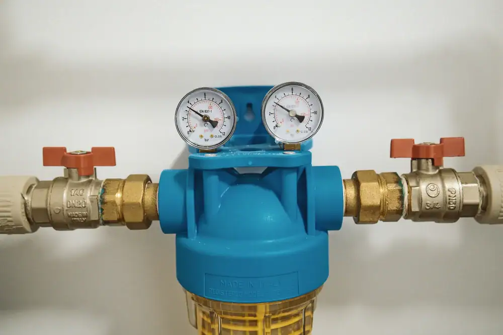 A close-up of a blue water filter system with two pressure gauges on top and two brass valves with red handles on either side, mounted against a white wall.