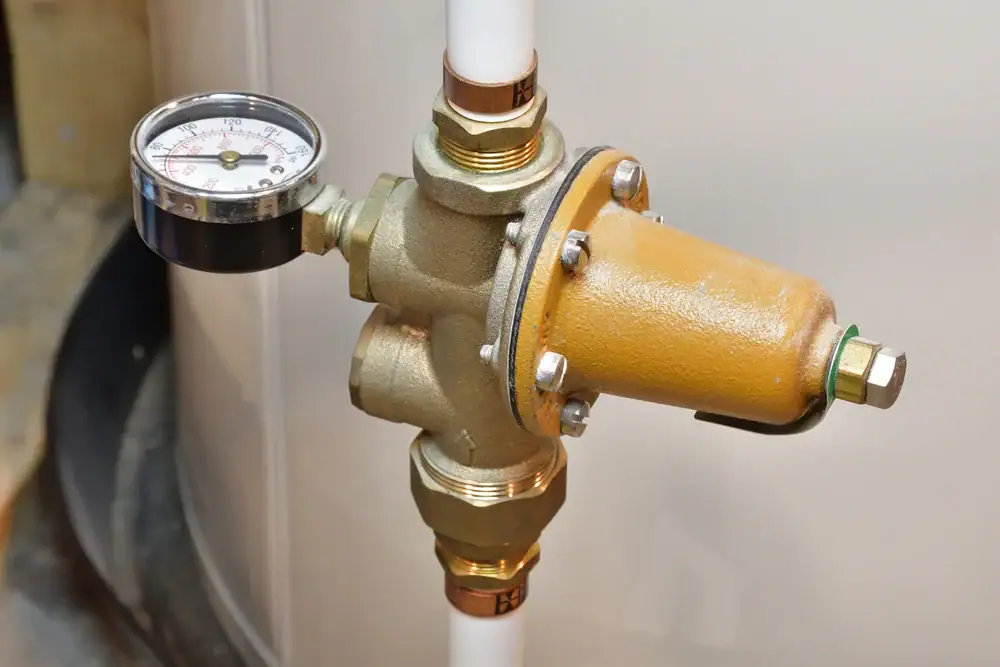A brass water pressure regulator with a gauge attached to white pipes, mounted against a light-colored wall. The gauge displays pressure in psi and kPa.