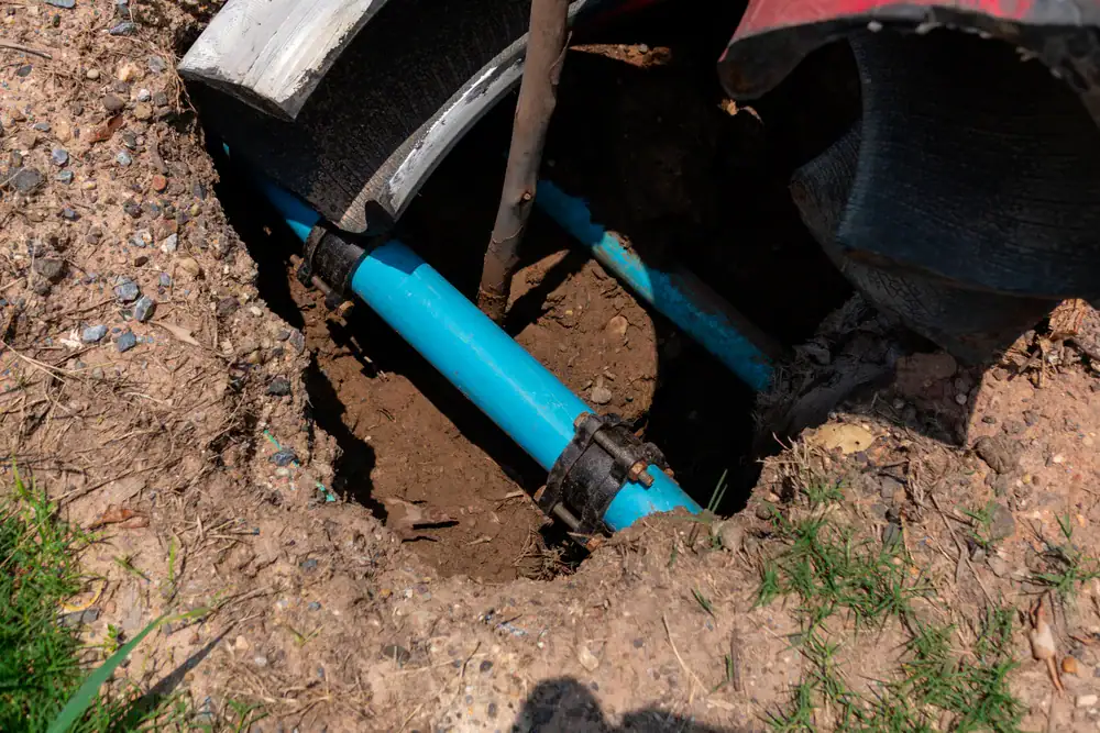 A blue water pipe with black connectors is exposed in a hole dug in the ground, surrounded by dirt, grass, and protective coverings.