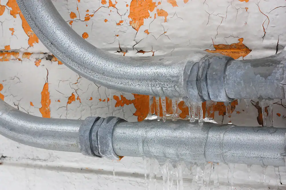Two metal pipes covered in ice are mounted on a wall with peeling white and orange paint. Icicles hang from the underside of the pipes, indicating freezing temperatures.