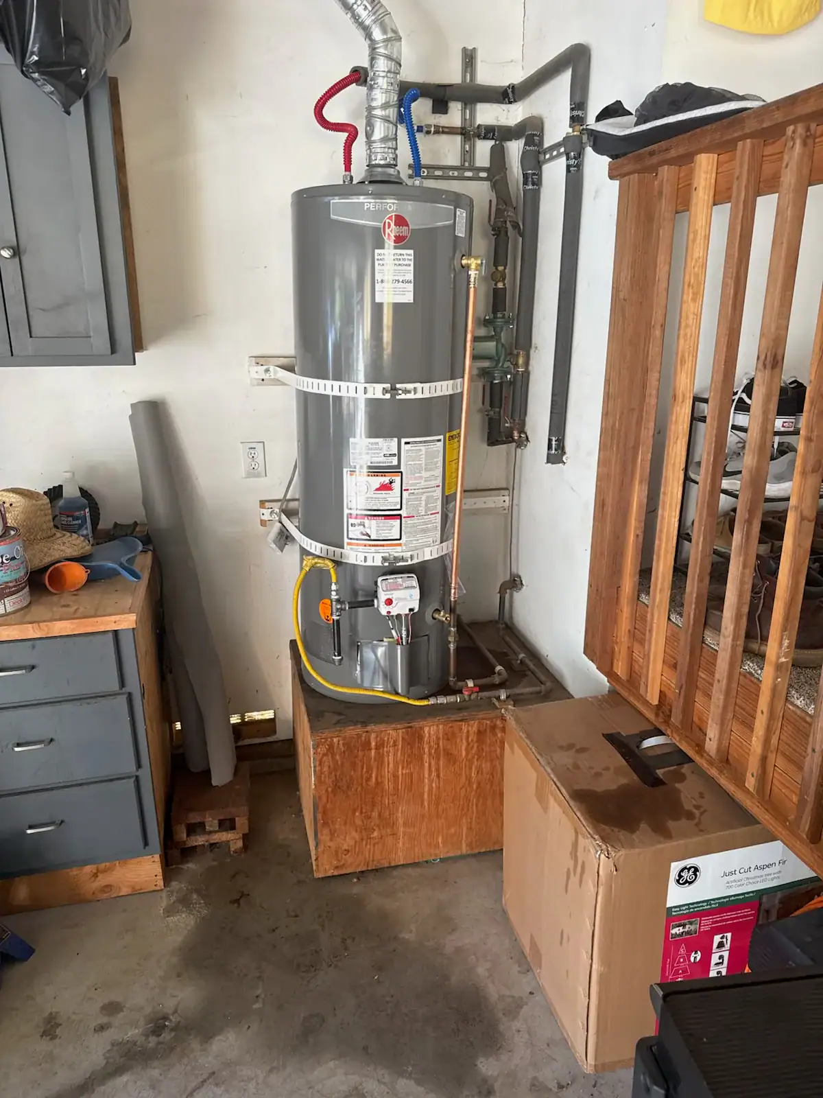 A gray water heater is installed in a corner of a garage, with pipes connected to the top. Nearby are a cardboard box, a wooden platform, and a workbench with tools and items on it.