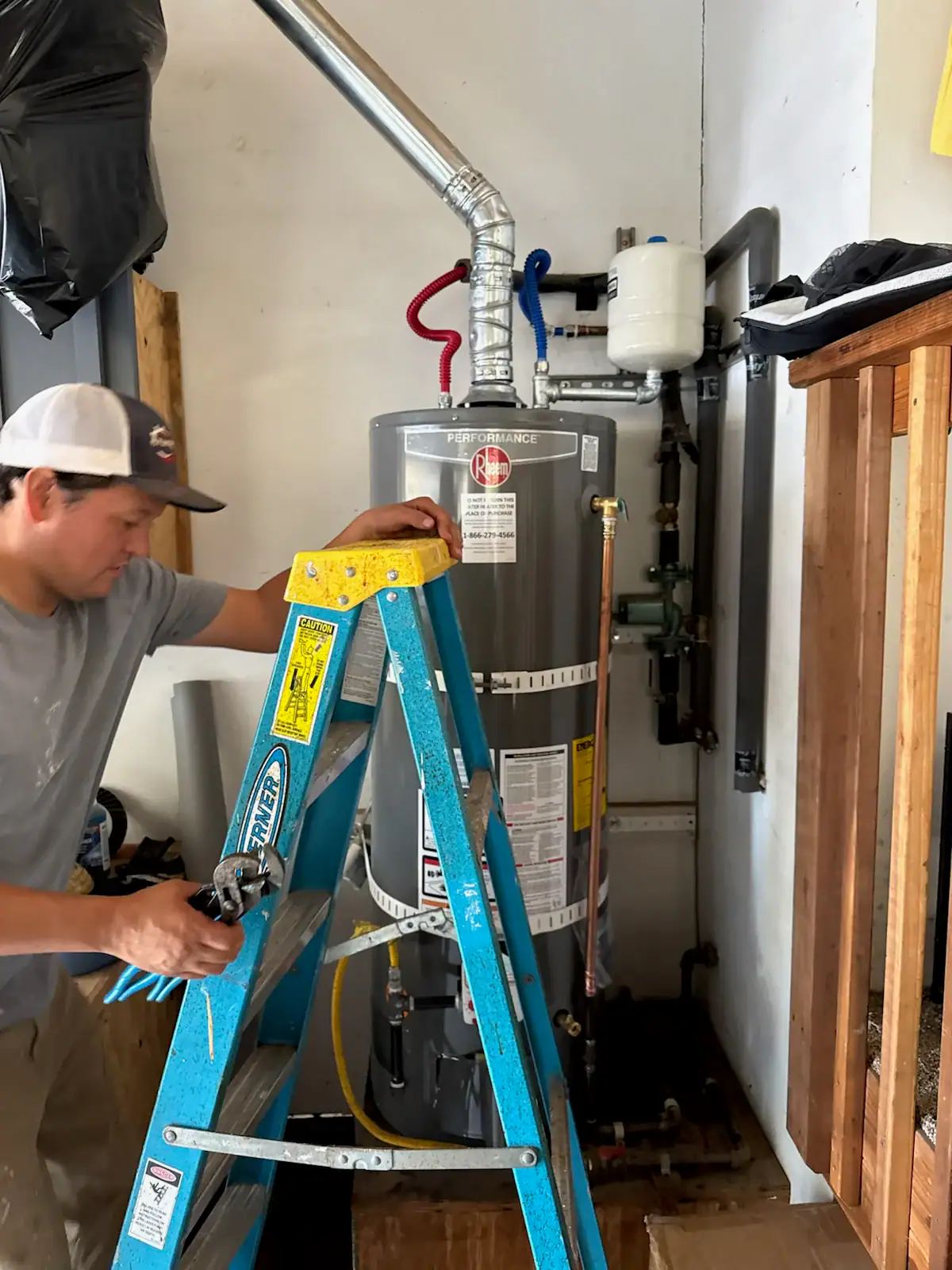 A man stands on a blue ladder working on a gray water heater in a utility room, using a tool. Pipes are connected to the water heater, and a wooden structure is visible on the right side.