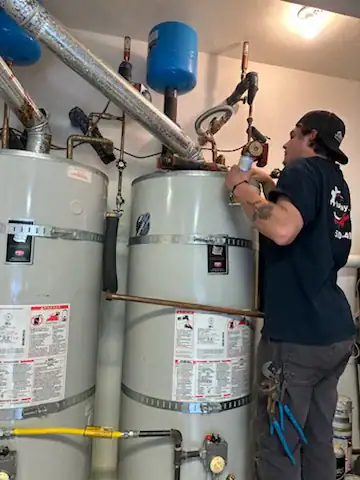 A technician wearing a black shirt and cap works on a set of large water heaters with various pipes and gauges in a utility room. Tools are attached to his belt, and he uses a wrench on one of the water heaters.