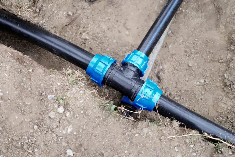 Three black plastic pipes connected with blue fittings are shown laid out in a T-shape and partially buried in the ground, surrounded by soil.