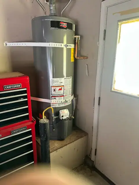 A gray Ruud water heater is installed on a raised platform in a garage next to a red Craftsman tool chest and a door with a window. The water heater is secured with white straps.